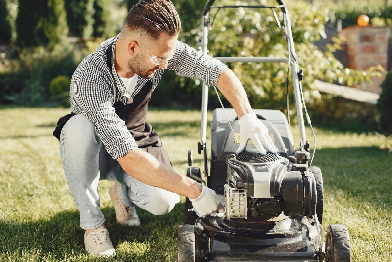 Man cutting grass with lawn mover in the back yard. Male in a black apron. Guy repairs.<br />